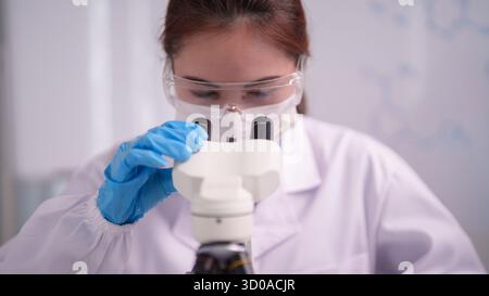 Eine engagierte Erwachsene asiatische Wissenschaftlerin untersucht Proben präzise mit einem Mikroskop. Sie trägt Sicherheitsausrüstung in einer hellen Laborumgebung. Stockfoto