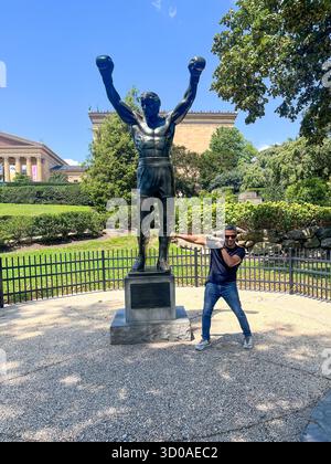 Ein Mann posiert vor einer Statue von Rocky. Die Statue zeigt einen Boxer mit erhobenen Armen. Der Mann trägt ein schwarzes Hemd und Jeans Stockfoto