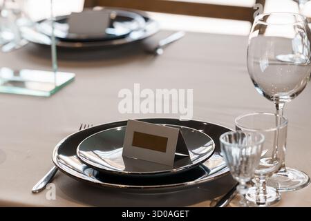 Der elegante Esstisch ist mit dunkelgrauer Bettwäsche, silberfarbenen Tellern, Weingläsern und einem Tischkartenhalter ausgestattet, was ein gehobenes Ambiente andeutet Stockfoto