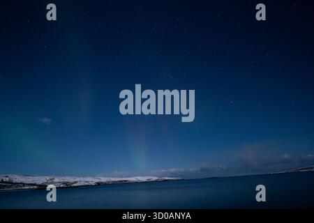 Breite horizontale Aufnahme eines tiefblauen Nachthimmels mit einem schwachen roten und grünen Schein der Aurora Borealis über einem ruhigen Gewässer und schneebedeckten Land Stockfoto