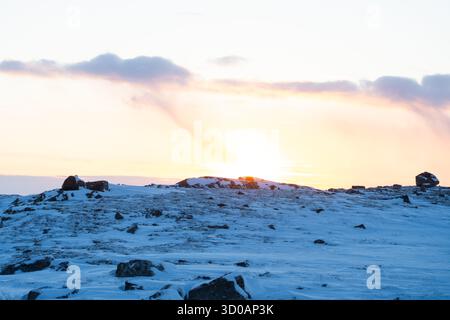 Winterlandschaft mit Sonnenuntergang hinter einem schneebedeckten felsigen Hügel mit weichen pastellfarbenen Wolken am Himmel Stockfoto