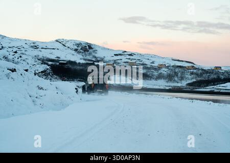 Murmansk, Russland - 13. Dezember 2024: Jetour T2 SUV fährt auf einer verschneiten Küstenstraße in Richtung arktischer Hügel. Dreiviertelansicht des SUV-Fahrens hinten Stockfoto