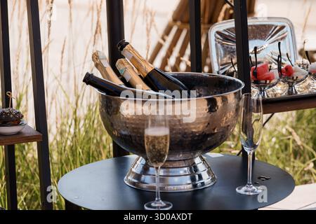 Ein elegantes Champagner-Ambiente im Freien verfügt über einen großen silbernen Eiskübel mit Flaschen, Kristallflöten und ein Erfrischungstablett, das an eine raffinierte Atmosphäre erinnert Stockfoto