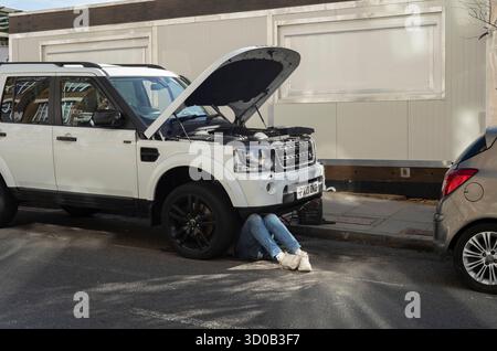 Ein Paar Beine eines Mechanikers, der unter dem Motor eines Land Rover arbeitet, der auf einer Wohnstraße in Hackney Wick, East London, Großbritannien, geparkt ist Stockfoto