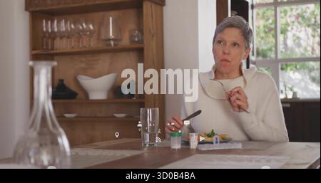 Mit Lesebrille und Gabel, Seniorin hält mitten am Küchentisch inne, mit Pillenflaschen Stockfoto