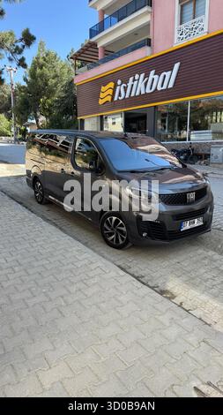27. Juni 2025, Antalya, Türkei. Ein Fiat-Van steht an einer asphaltierten Straße, mit einem Café und Bäumen im Hintergrund unter klarem Himmel. Stockfoto
