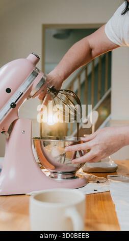 Eine Frau benutzt einen rosa Kitchenaid-Mixer, um einen Kuchen zu machen. Sie hält eine Schüssel mit einem Teig drin und ist dabei, ein paar Schokoladenchips hinzuzufügen Stockfoto