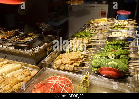 Auf einem geschäftigen Markt im Freien werden verschiedene Spieße mit frischem Gemüse und mariniertem Fleisch präsentiert. Die untergehende Sonne strahlt ein warmes Leuchten aus, enha Stockfoto