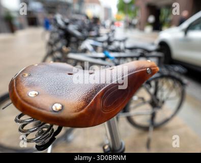 Nahaufnahme eines Fahrradsattels aus Vintage-Leder, nass von Regen, mit verschwommener Stadtstraße und geparkten Fahrrädern im Hintergrund. Stockfoto