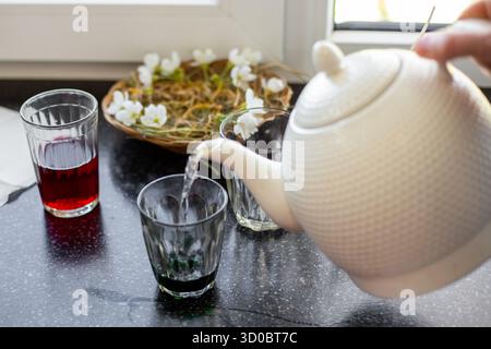 Eine Hand gießt heißes Wasser aus einer weißen Teekanne in ein Glas mit Farbstoff, um Ostereier zu färben. Stockfoto
