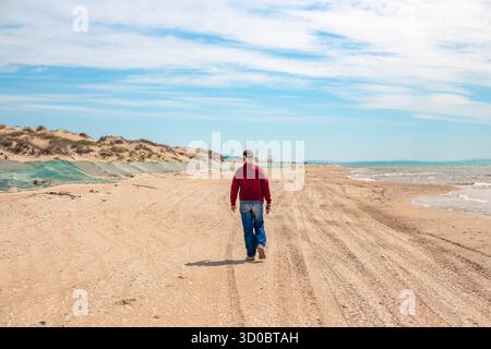 Ein Mann läuft an einem sonnigen Tag an einem Sandstrand entlang. Entlang des Meeres ist ein Sanddamm mit einem Netz bedeckt, um Heizöl zu fangen. Stockfoto