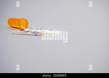 Eine verschreibungspflichtige Flasche mit kleinen weißen Pillen wird auf einem 100-Dollar-Schein verschüttet. Wird auf grauem Hintergrund von einem Ebenen Blickpunkt aus betrachtet. Stockfoto