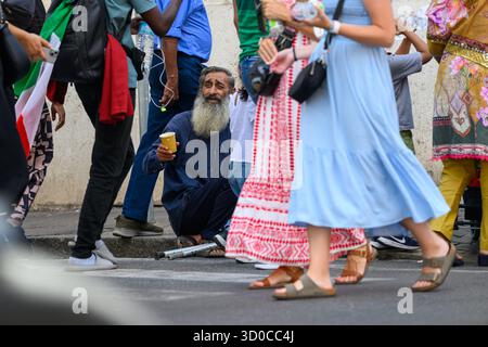 Ein bärtiger, älterer Mann sitzt auf dem Bordstein und bettelt um Geld, während eine geschäftige Menschenmenge vorbeizieht. Via di Porta Angelica, Rom, Italien. Stockfoto