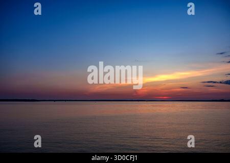 Ruhige Sonnenuntergangsszene mit warmem Orange und rosa Licht, das sich auf stillem Wasser reflektiert, und einem blauen Himmel mit Farbverlauf über dem Horizont Stockfoto