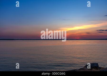 Ruhige Sonnenuntergangsszene mit warmem Orange und rosa Licht, das sich auf stillem Wasser reflektiert, und einem blauen Himmel mit Farbverlauf über dem Horizont Stockfoto
