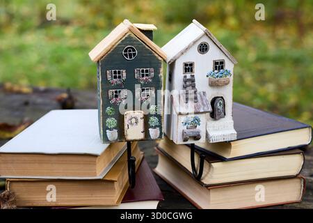Nahaufnahme von zwei dekorativen Holzhäusern, die auf Stapeln von Büchern platziert sind, vor einem natürlichen grünen Hintergrund, wodurch eine skurrile Szene entsteht. Stockfoto