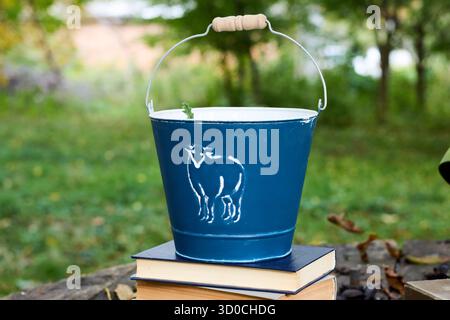 Vintage-blauer Eimer mit weißem Schafdesign, der draußen auf einem Stapel Bücher sitzt und eine nostalgische und skurrile Szene schafft Stockfoto