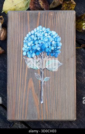 Handgemalte blaue Hortensien auf braunem Holzbrett, umgeben von trockenen Blättern und Nüssen auf einem rustikalen Holztisch, kunstvolle Blumendekoration Stockfoto