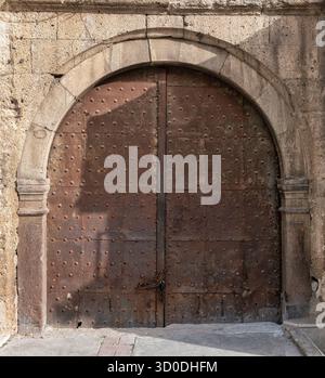 Alte Bogentür aus rostigem, nietenbesetztem Metall, gesichert durch eine Kette und ein Vorhängeschloss, in einem verwitterten Steinbogen und einer rauen Mauer. Stockfoto