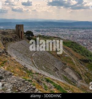 Panoramablick auf das steile antike griechische Theater der Pergamon-Ruinen, mit der Stadt Bergama, der Türkei und den fernen Bergen unter einem bewölkten Himmel. Stockfoto