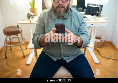 Mann, der drinnen auf einem Stuhl sitzt und ein Smartphone benutzt. Stockfoto