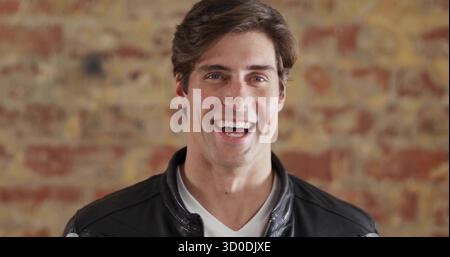 Ein lächelnder Mann, der eine Lederjacke trägt, posiert vor einer Ziegelwand, mit warmer Beleuchtung Stockfoto
