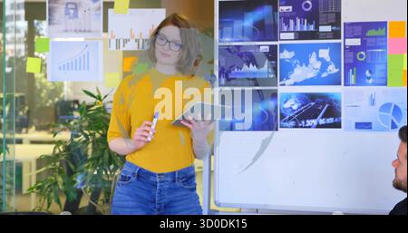 Frau im Senfpullover, der im Büro auf das Whiteboard zeigt, mit Tablet und Haftnotizen Stockfoto