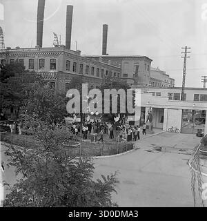 Eine aus der Luft gezogene Hochwinkelansicht des Haupteingangs (prokhodnaya) der Armature-Isolator-Anlage (AIZ) in Sloviansk, 1970er Jahre Dieses Archivfoto aus der UdSSR, der ukrainischen SSR, zeigt eine große Menge von Arbeitern, die sich auf dem Fabrikplatz zu einer Feier versammelt haben (Ballons sind sichtbar). Der Hintergrund wird von den massiven, historischen Backsteingebäuden (Tsekh) mit hohen Schornsteinen dominiert. Dieses Bild fängt die klassische sowjetische Industrielandschaft und die Fabrik als Zentrum des gesellschaftlichen Lebens ein. Ein wertvolles Dokument über das friedliche industrielle Leben im Donbass vor dem Krieg Stockfoto