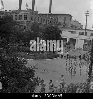 Eine aus der Luft gezogene Hochwinkelansicht des Haupteingangs (prokhodnaya) der Armature-Isolator-Anlage (AIZ) in Sloviansk, 1970er Jahre Dieses Archivfoto aus der UdSSR, der ukrainischen SSR, zeigt eine große Menge von Arbeitern, die sich auf dem Fabrikplatz zu einer Feier versammelt haben (Ballons sind sichtbar). Der Hintergrund wird von den massiven, historischen Backsteingebäuden (Tsekh) mit hohen Schornsteinen dominiert. Dieses Bild fängt die klassische sowjetische Industrielandschaft und die Fabrik als Zentrum des gesellschaftlichen Lebens ein. Ein wertvolles Dokument über das friedliche industrielle Leben im Donbass vor dem Krieg Stockfoto