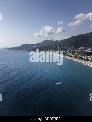 Blick aus der Vogelperspektive auf das türkisfarbene Wasser trifft auf die Sandküste, die von üppigen grünen Hügeln und der Stadtlandschaft von Alanya, Antalya, Türkei, umgeben ist. Stockfoto