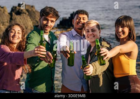 Eine fröhliche, multiethnische Gruppe von fünf jungen Freunden blickt direkt in die Kamera, während sie einen feierlichen Toast mit grünen Bierflaschen an der se macht Stockfoto