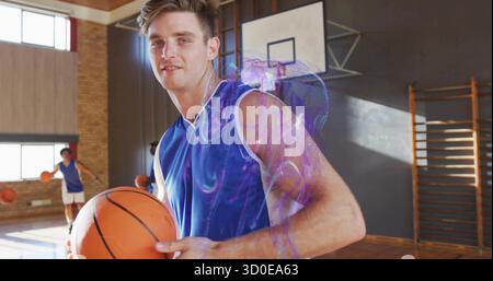 Stehender Mann in blauem Jersey, der Basketball in der Turnhalle hält, mit Teamkollegen Dribbling Ball in der Nähe des Basketballkörpers Stockfoto