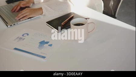 Ein erwachsener Mitarbeiter kann am Schreibtisch mit einem silbernen Laptop arbeiten, mit ausgedrucktem Bericht und Kaffeetasse Stockfoto