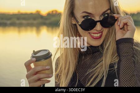 Closeup Outdoor Portrait von niedlichen Happy blond Kaukasischen erwachsenen Frau in glücklichen Zustand mit Kaffee zum Mitnehmen. Junge Frau mit langem Haar Sonnenbrille tragen Stockfoto