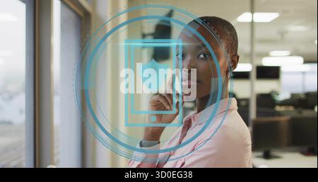 Stehende Frau in rosa Hemd, die neben einem großen Fenster im Großraumbüro blickt, mit digitaler Überlagerung Stockfoto