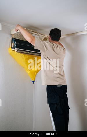 Vertikaler Schuss des HLK-Technikers, der die Klimaanlage vorsichtig demontiert und dabei tropfendes Wasser und Fremdkörper während der Reparatur mit einem Schutzbeutel aufgefangen Stockfoto