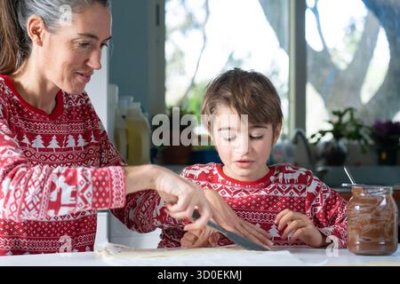 Mutter und Sohn tragen festliche Pyjamas und verteilen Schokolade auf Teig in der Küche Stockfoto