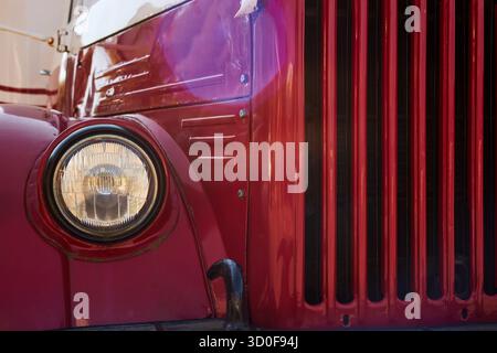 Nahaufnahme mit dem vorderen Teil des roten Vintage-Trucks mit rundem Scheinwerfer und vertikalem Kühlergrill, der klassische Details des Automobildesigns und das reflektierende Sonnenlicht auf der Metalloberfläche erfasst Stockfoto