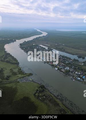 Aus der Vogelperspektive auf einen sich windenden Fluss, der den weichen, bewölkten Himmel reflektiert, während er durch ein Dorf inmitten üppiger grüner Feuchtgebiete in Bukarest, Rumänien, schneidet. Stockfoto