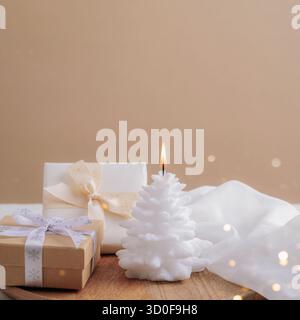 Festliche weiße brennende Kerze, Geschenkboxen auf beigefarbenem Hintergrund mit Weihnachtslichtern. Nahaufnahme, Kopierraum. Stockfoto