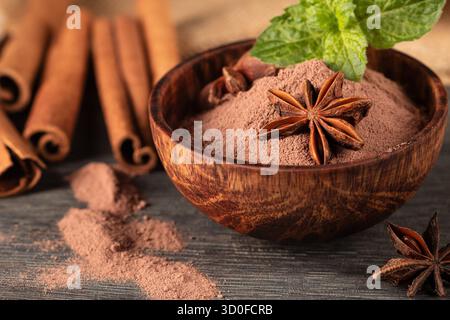 Diese Nahaufnahme zeigt eine rustikale Holztasse gefüllt mit reichhaltigem Kakaopulver, begleitet von duftendem Sternanis und warmen Zimtstäbchen, die kreieren Stockfoto