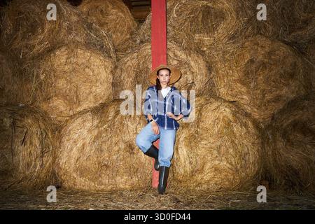 Porträt einer jungen erwachsenen Kaukasierin mit Strohhut, der sich vor gestapelten Heuballen an einen roten Holzpfosten lehnt, mit einem Bein gebeugt und die Hände auf den Hüften gelegt Stockfoto