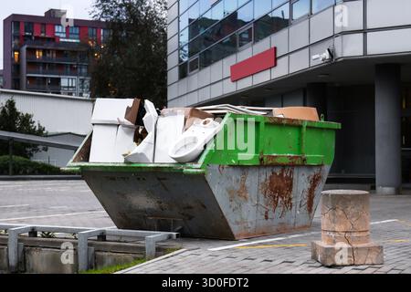 Ein grüner Behälter für Bauabfälle, gefüllt mit Abfällen, einschließlich alter Möbel und Geräte, befindet sich außerhalb eines modernen Gebäudes. Stockfoto