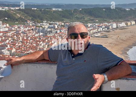 Ein Mann mittleren Alters mit dunkler Sonnenbrille und grauen Haaren posiert im Vordergrund mit der malerischen Küstenstadt Nazare, Portugal, dem Strand Stockfoto