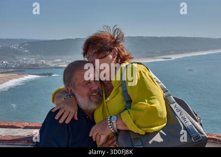 Ein älteres Ehepaar posiert zärtlich umarmt an einem Aussichtspunkt in Nazare, Portugal, mit dem Atlantik und der Küste im Hintergrund. Stockfoto