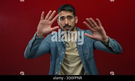 Junger hispanischer Mann in lässiger Kleidung posiert selbstbewusst vor einem leuchtend roten Hintergrund und drückt verschiedene Emotionen mit dynamischen Handgesten aus. Stockfoto