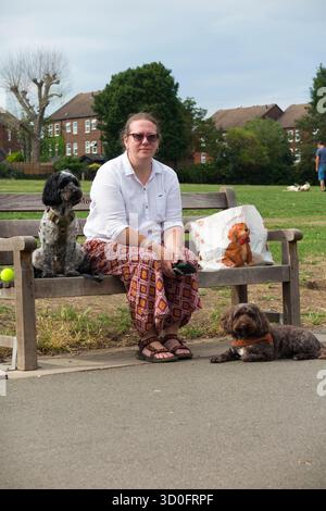 Hundeschlittenfahrerin, Hundeliebhaberin/Hundeliebhaberin macht eine Pause, setzt sich auf eine Parkbank, während eines Spaziergangs mit ihren beiden Hunden und ihrer Einkaufstasche mit dem Thema Hund. Kneller Gardens in Twickenham. UK. (145) Stockfoto