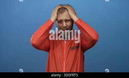 Mann im orangefarbenen Gefangenenanzug, der Kopf mit beiden Händen hält, in einem Studio-blauen Setting, Häftlingszahl sichtbar; Angst. Stockfoto