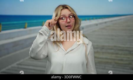 Frau mit blonden Haaren und Brillen, die auf einer sonnigen Promenade am blauen Meer spielerische Gesten macht, in weiß, was an eine entspannte und fröhliche Küste erinnert Stockfoto