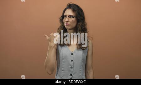 Frau in ärmelloser Weste und Brille zeigt im Studio den Daumen zur Seite, während sie vor der Kamera runzelt; Ekel. Stockfoto
